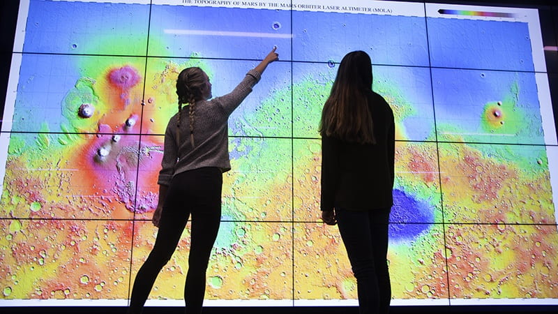 Image is of a large dataset being dislplayed in the Price Science Library's Visualization Lab, which is a series of 16 high definition screens. Two people are looking at the screen, and appear as silhouettes/backlit.